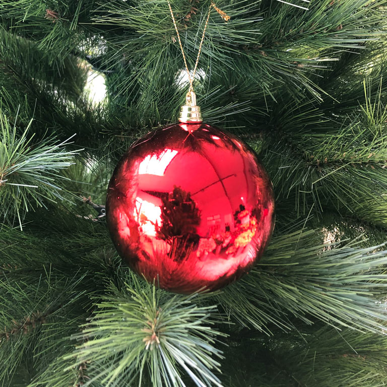 A red, round, shiny Christmas ornament hanging on a Christmas tree with green needles.