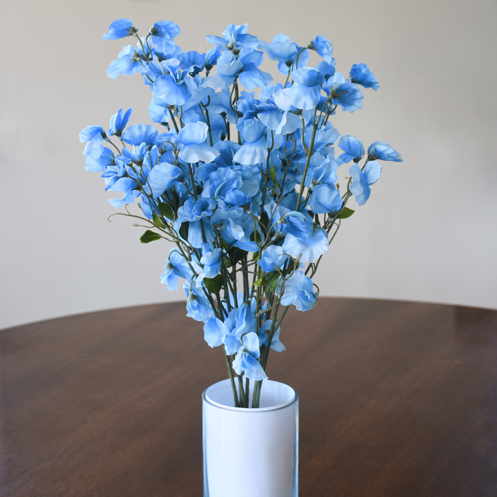 A bouquet of artificial light blue flowers with green stems arranged in a white cylindrical vase on a dark wooden table against a plain beige wall.