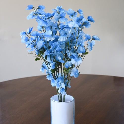 A bouquet of artificial light blue flowers with green stems arranged in a white cylindrical vase on a dark wooden table against a plain beige wall.