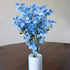 A bouquet of artificial light blue flowers with green stems arranged in a white cylindrical vase on a dark wooden table against a plain beige wall.